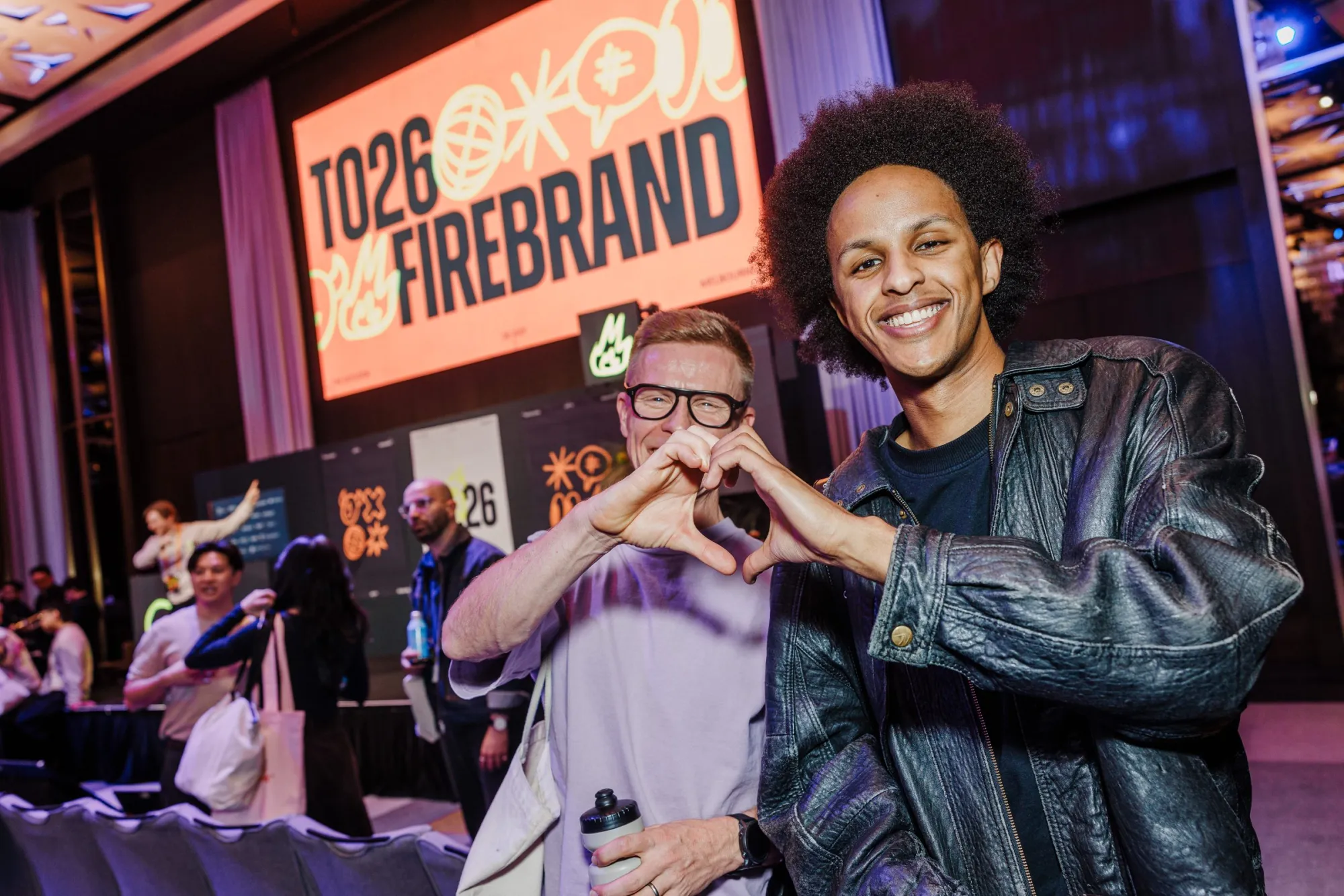 Two attendees make a heart with their hands in front of the TO26: Firebrand stage screen.